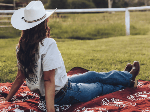 Red and white patterned recycled picnic blanket spread on grass with person wearing white cowboy hat seated on it.