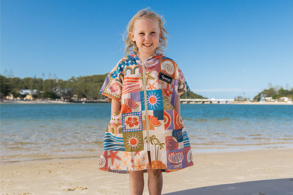 Kids hooded towel in multicolored retro beach print with geometric sea motifs, worn by child on sandy beach.