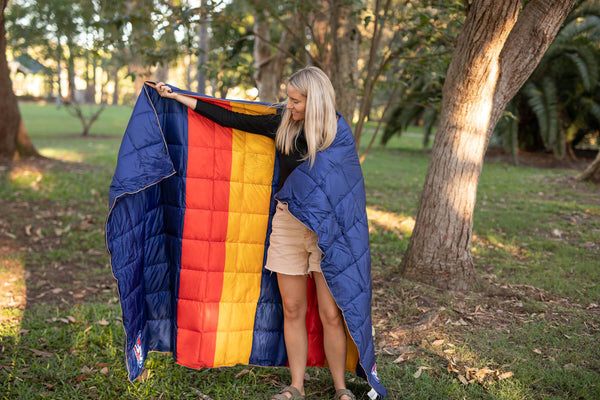 Navy puffy blanket with red and gold central stripe, worn draped around person in outdoor setting, showing quilted texture an