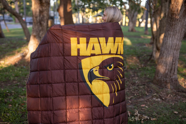 Maroon puffy blanket with yellow "HAWKS" text and eagle logo, worn draped over person's shoulders outdoors.