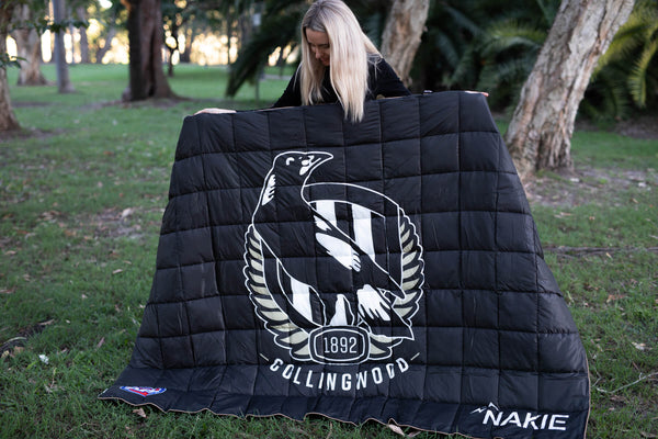 Black quilted puffy blanket with Collingwood Magpies eagle logo and 1892 crest, displayed outdoors by woman with blonde hair