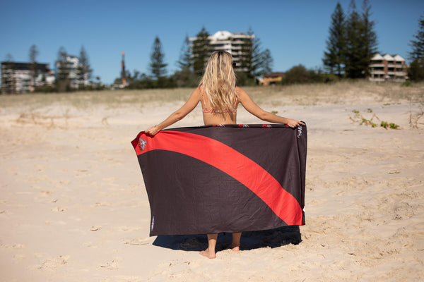Black and red beach towel with diagonal stripe design, displayed on sand by person facing away from camera