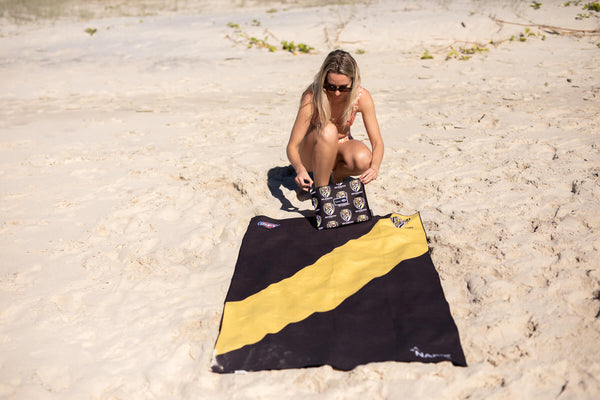 Black and gold Richmond Tigers AFL beach towel with diagonal stripe design, displayed on sand at the beach