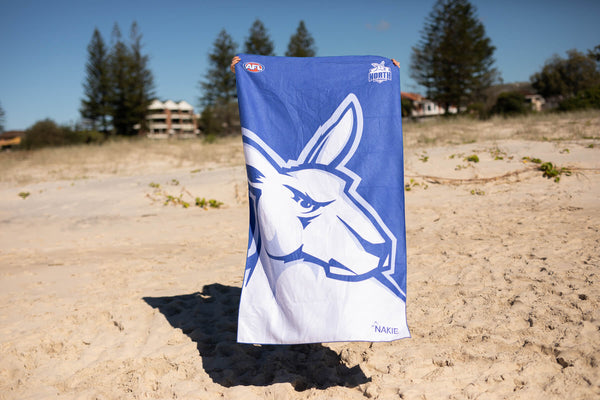 North Melbourne Kangaroos blue beach towel with white kangaroo logo and AFL branding, displayed on sand.