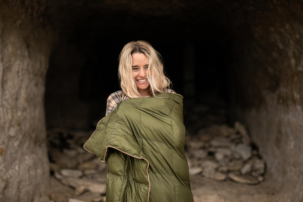 Olive green down puffy blanket wrapped around woman's shoulders, quilted design with tan trim.
