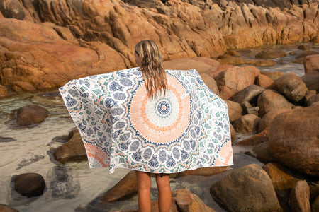 Woman holding Nakie's recycled sand-free beach towel on her back while facing a natural rock pool, enjoying sustainable comfort amidst the serene natural surroundings.
