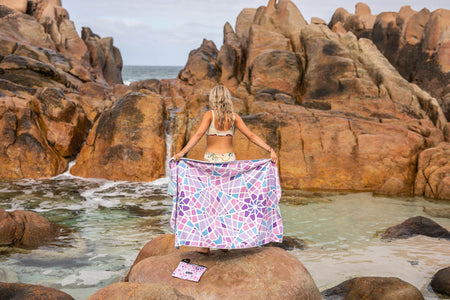 Woman holding Nakie's recycled sand-free beach towel on her back while facing a natural rock pool, enjoying sustainable comfort amidst natural beauty.