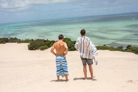 Image of two men wrapped in Nakie's recycled sand-free beach towel standing on a beach, admiring the view and enjoying sustainable comfort during their beach outing.