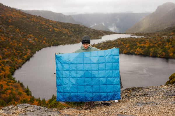 Blue quilted down puffy blanket held up outdoors with mountain lake landscape behind