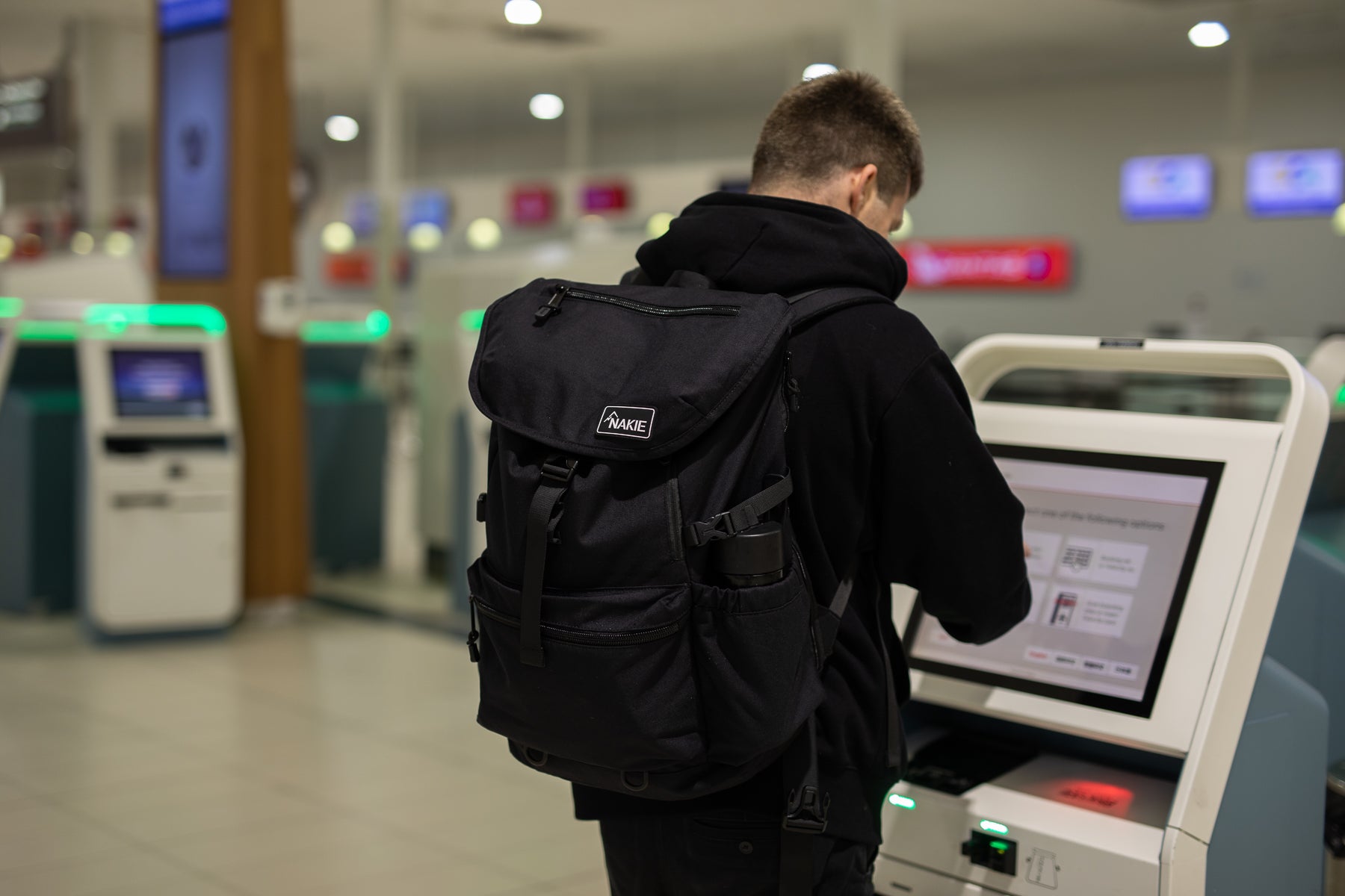 Person using a self-check-in kiosk at the airport while wearing a Nakie Midnight Black travel backpack.