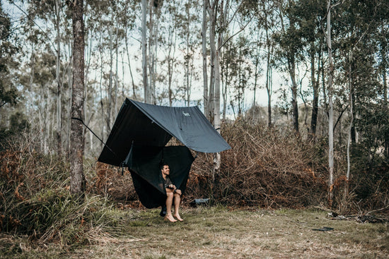 One person standing under a Nakie hammock, bug net, and rain tarp setup in a sparse forest.