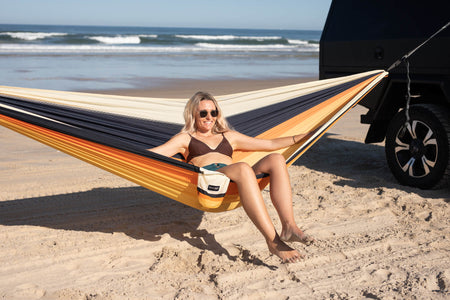 Striped camping hammock in cream, navy, and gold with woman relaxing on beach near vehicle and ocean waves