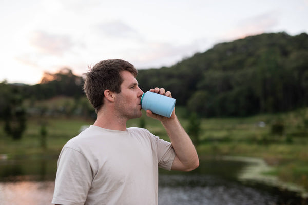 Light blue insulated coffee cup with lid, held by man drinking outdoors near water and green hills