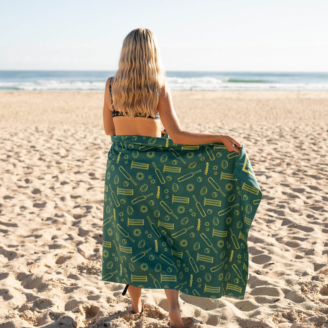 Green recycled beach towel with yellow cricket and summer pattern, shown draped on person at beach from behind.