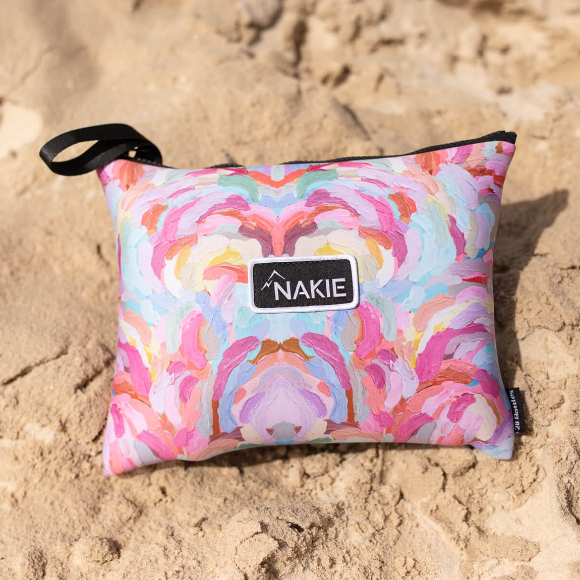 Colorful geometric patterned beach towel in pink, orange, yellow and blue with black handle strap on sand.