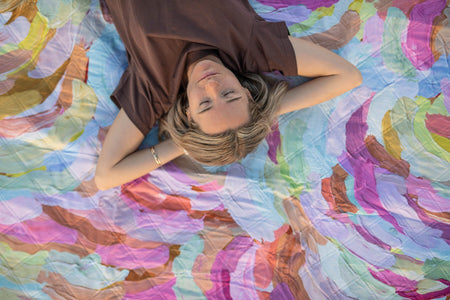 Recycled picnic blanket in multicolor watercolor floral print, displayed overhead with woman lying on it.
