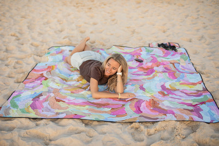 Recycled picnic blanket with multicolored swirl pattern in pink, purple, blue, and green laid out on sand.