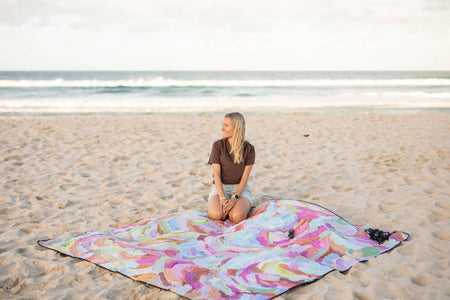 Recycled picnic blanket with multicolored abstract watercolor print, spread on beach sand with woman kneeling on it
