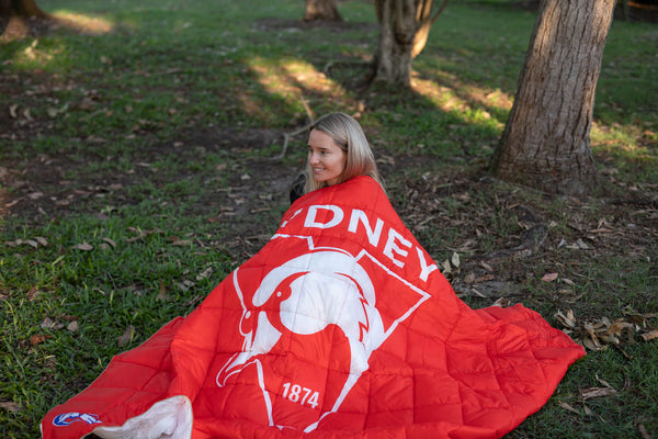 Red and white Sydney Swans AFL puffy blanket with logo and "1874" text, draped over person outdoors