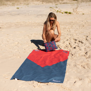 Red and navy colorblock beach towel with AFL branding, displayed on sand with matching patterned pouch.