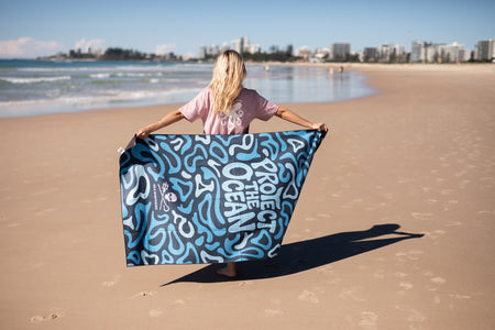 Recycled sand-free beach towel with blue abstract wave pattern, displayed on sandy beach by ocean.