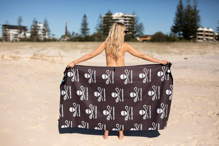 Black recycled beach towel with white skull and utensils print, held open on sandy beach