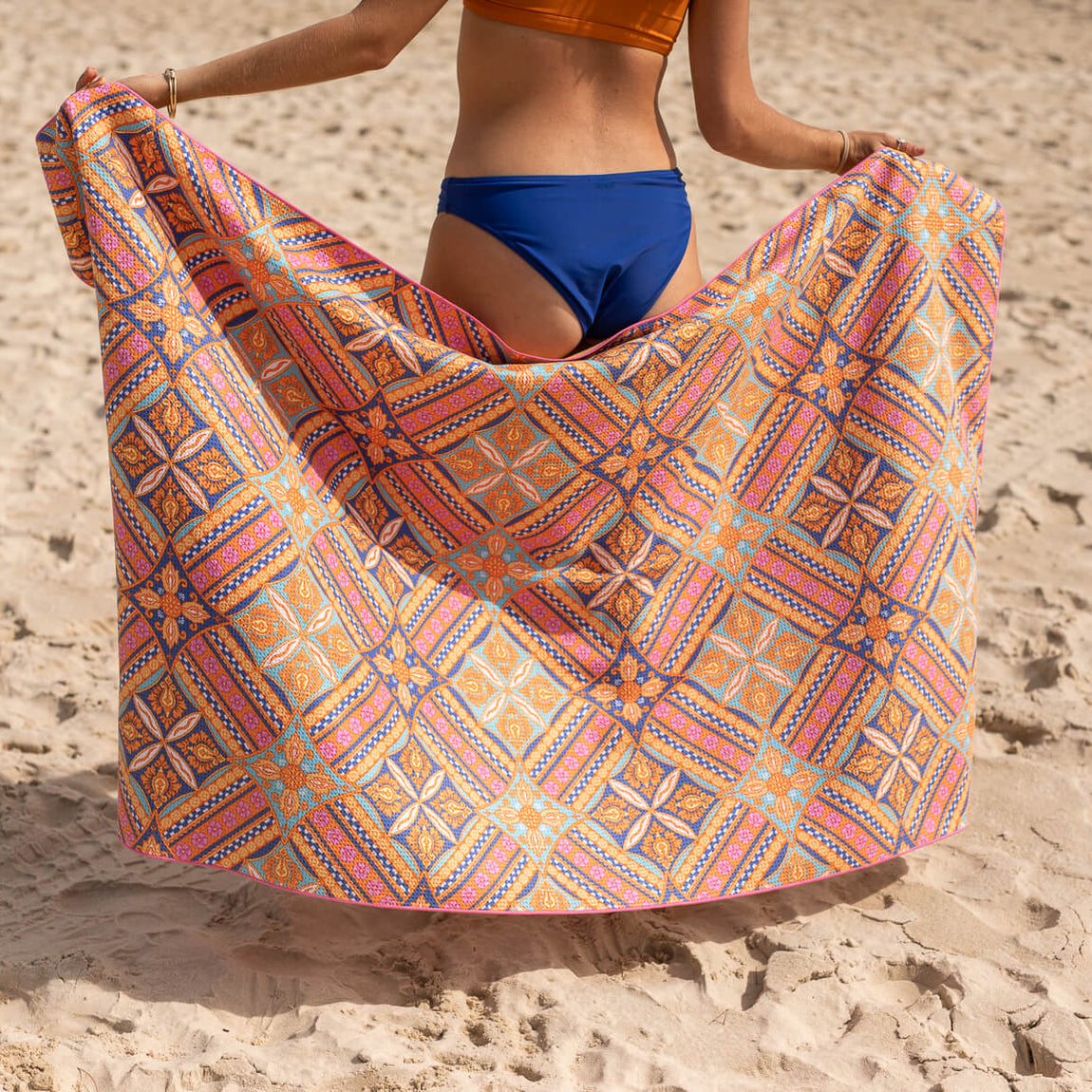 Multicolored geometric patterned beach towel in orange, pink, and blue tones, displayed on sand.