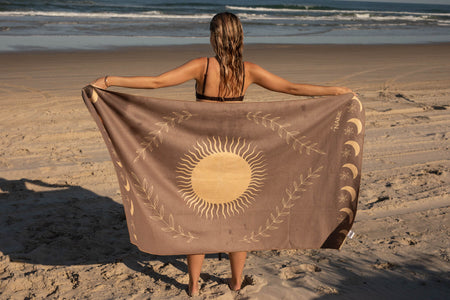 Beige recycled beach towel with gold sun and celestial design, held open on sandy beach