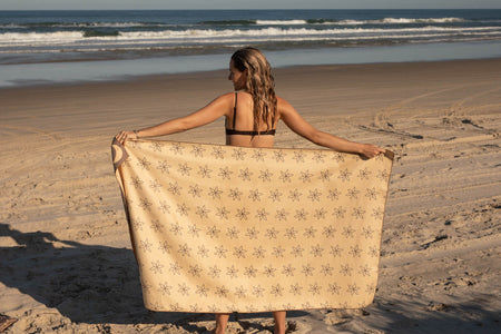 Cream beach towel with all-over star pattern, held open by woman in bikini on sandy beach with ocean waves behind