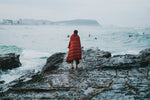 Image of a man wrapped in Nakie's Earth Red Sustainable Down Puffy Blanket, watching other people swim, enjoying warmth and sustainability by the water. preview #7