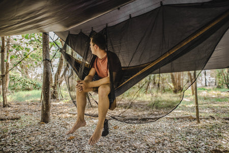 A man sitting comfortably in a Nakie hammock, protected by our Bug Net & Rain Tarp Combo, showcasing the ultimate outdoor relaxation experience with complete protection from insects and rain.