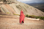 Image of a smiling woman wrapped in Nakie's Earth Red Sustainable Down Puffy Blanket, enjoying warmth and comfort during outdoor adventures. preview #10