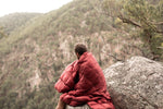Image of a man wrapped in Nakie's Earth Red Sustainable Down Puffy Blanket, enjoying the mountain view while sitting on a cliff, embracing warmth and sustainability during outdoor exploration. preview #9