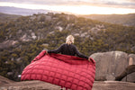 Image of a woman holding Nakie's Earth Red Sustainable Down Puffy Blanket behind her back, showcasing its vibrant color and sustainable design. preview #5